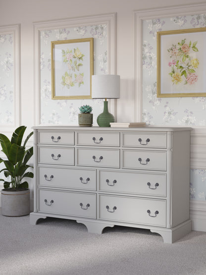 A dove grey Laura Ashley Clifton 6+4 drawer chest with traditional design against a floral background with a house plant next to it, a table lamp on top with various ornaments