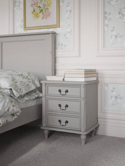 A dove grey 3-drawer bedside chest with traditional design and metal handles next to a bed with additional accessories