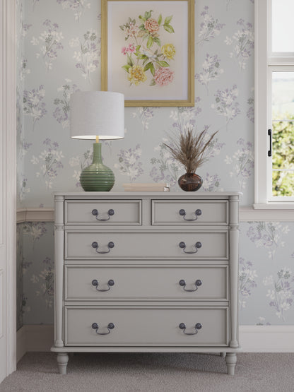 A dove grey chest of drawers with two smaller drawers on top and three larger ones below, each with a dark metal handle. Set against a floral wallpapered wall with a table lamp and plant on top.
