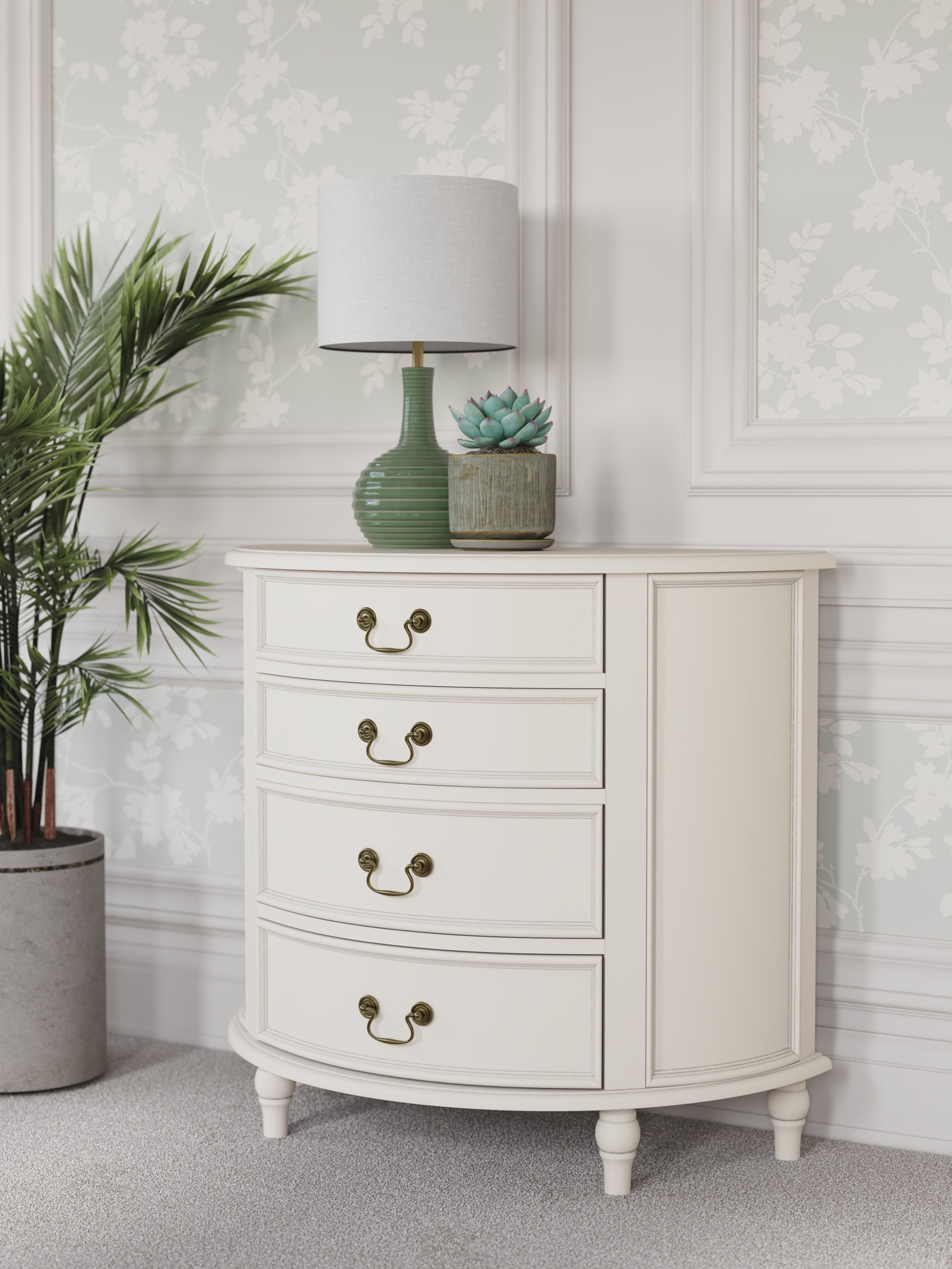 A left-hand corner view of an Ivory coloured half moon chest with four drawers and vintage styling. A lamp and some plants are shown with it