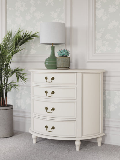 A left-hand corner view of an Ivory coloured half moon chest with four drawers and vintage styling. A lamp and some plants are shown with it