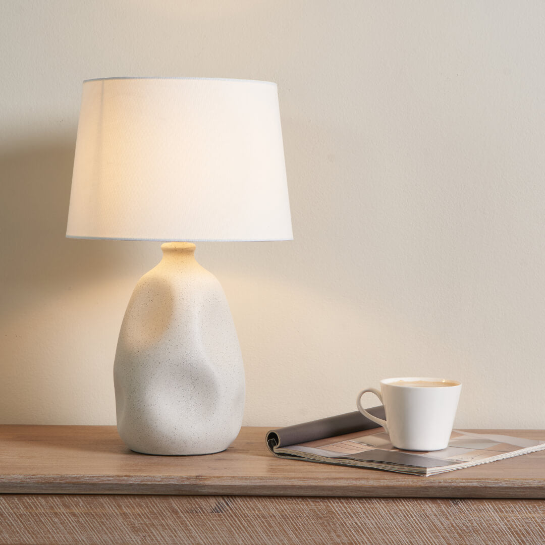 Table lamp with a white shade on a wooden surface next to a cup and magazine.